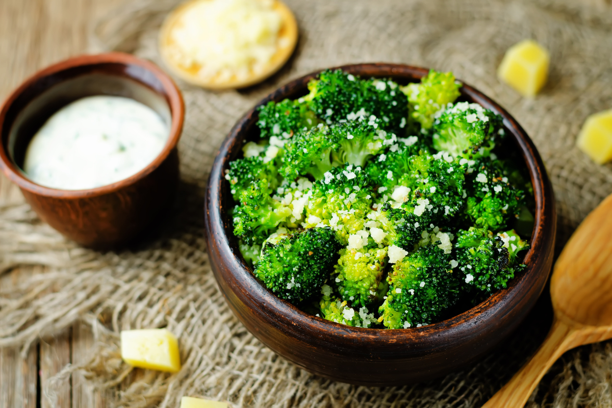 A rustic bowl filled with tender broccoli florets, lightly sprinkled with grated cheese. A creamy dipping sauce sits nearby for an extra savory touch