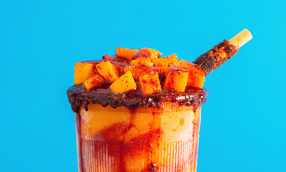 A vibrant mangonada, featuring fresh mango chunks drizzled with tangy chamoy sauce, topped with a tamarind candy stick, and beautifully presented against a bright blue background. Perfect for highlighting authentic Mexican flavors in your article.