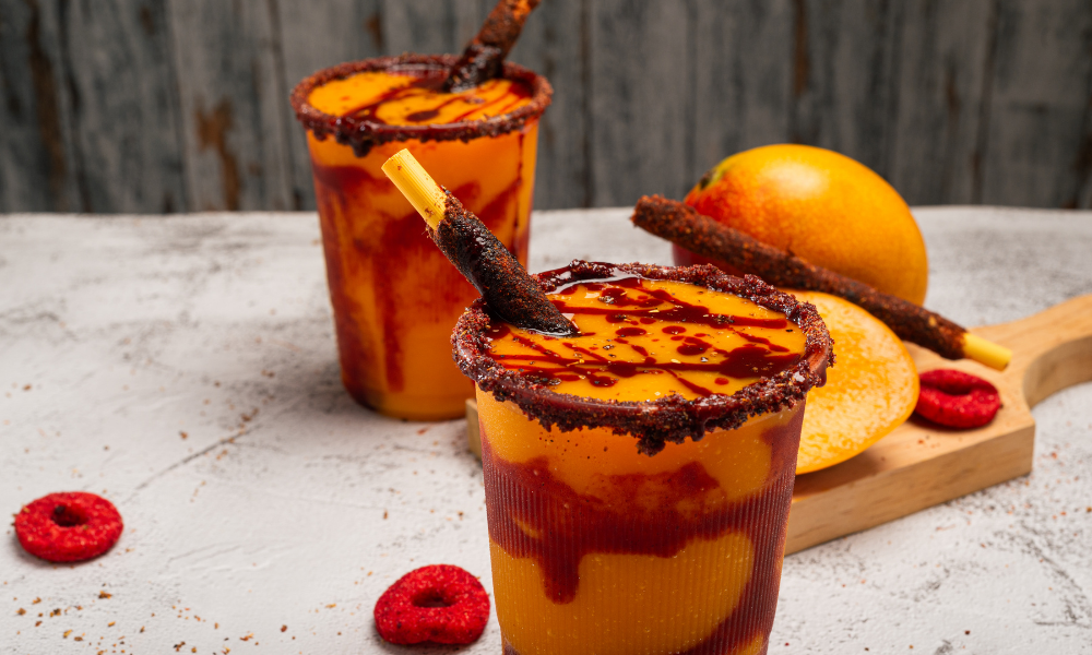 Two vibrant mangonadas in clear cups, featuring rich layers of mango puree and tangy tamarind sauce, rimmed with chili powder and topped with tamarind candy sticks. Fresh mango slices and raspberries complete this classic Mexican treat, perfect for any article showcasing authentic flavors.