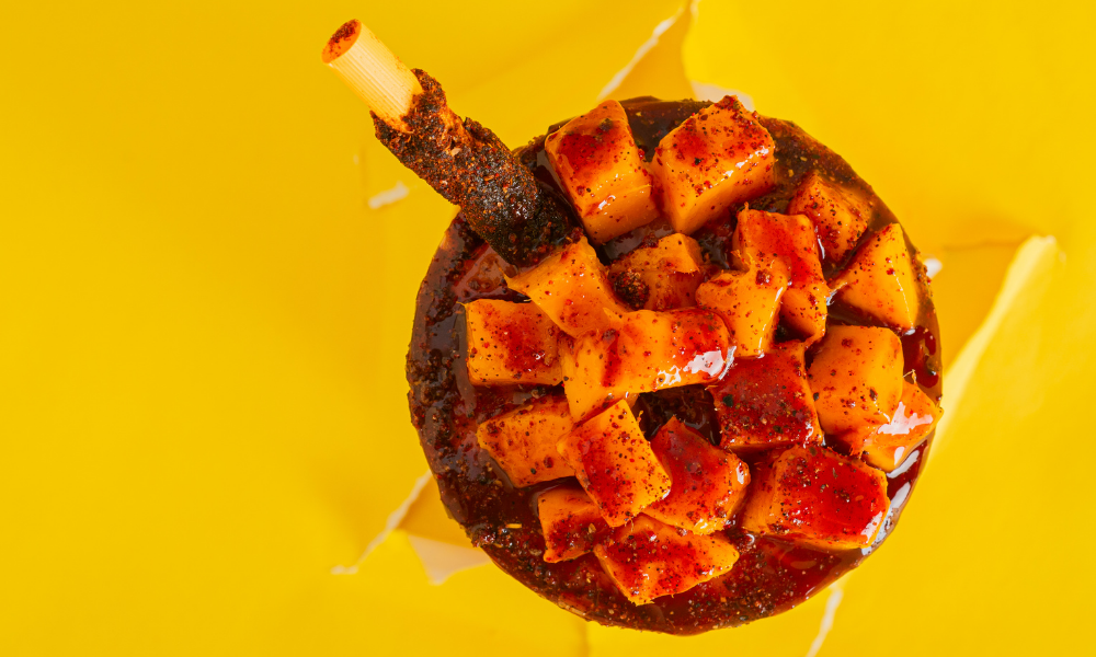 Top-down view of a mangonada bursting with vibrant mango chunks coated in tangy sweet and tangy sauce and sprinkled with chili powder, crowned with a tamarind candy stick. Set against a bold yellow background, this image highlights the bold and refreshing flavors of this iconic Mexican treat.