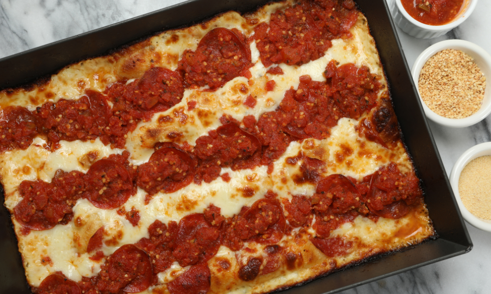 A freshly baked Detroit-style pizza in a rectangular pan, topped with layers of melted cheese, crispy pepperoni, and rich tomato sauce arranged in traditional stripes. The golden caramelized edges of the cheese give it a crispy, delicious crust.