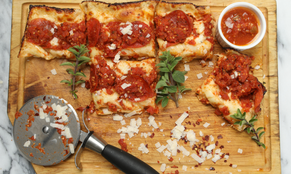 A serving of Detroit-style pizza on a wooden board, garnished with fresh oregano and parmesan shavings. A pizza cutter sits nearby, with a small bowl of dipping sauce and seasonings complementing the meal.