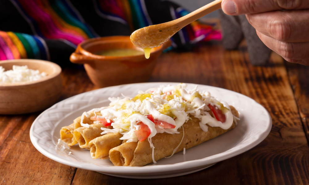 A plate of crispy flauta topped with sour cream, shredded lettuce, tomatoes, and grated cheese, being drizzled with sauce. 