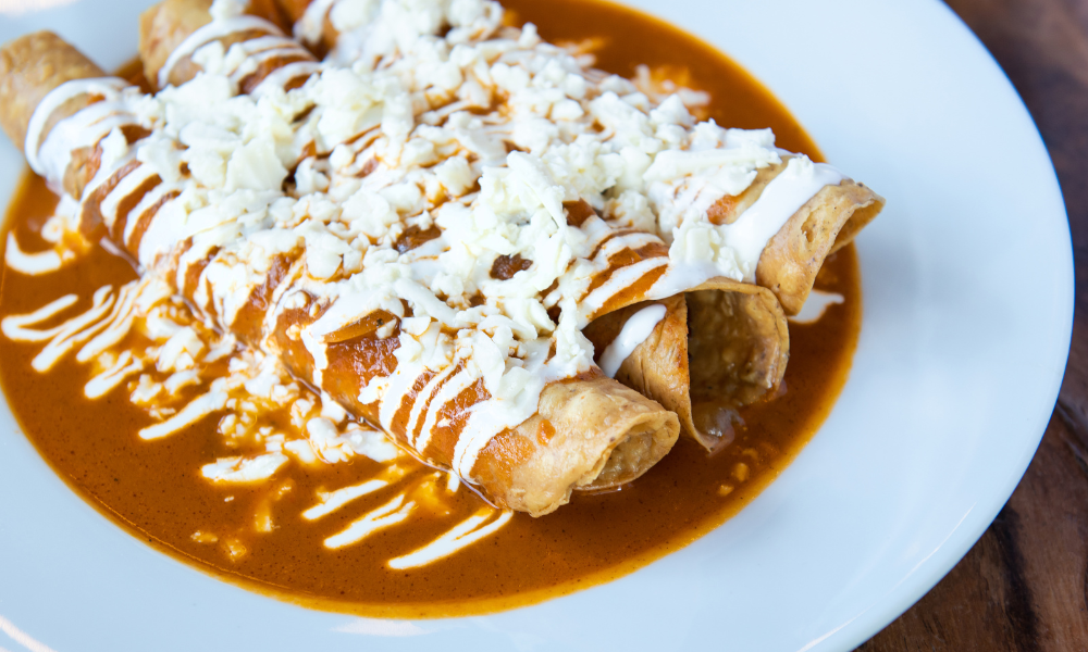 Crispy flauta served with a rich red sauce, sour cream drizzle, and crumbled cheese on a white plate