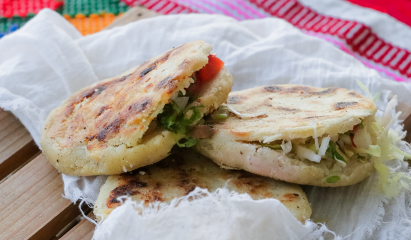 Three rustic gorditas filled with shredded lettuce, tomato, and other fresh ingredients, served on a white cloth atop a wooden surface. The colorful Mexican textiles in the background enhance the authentic presentation.