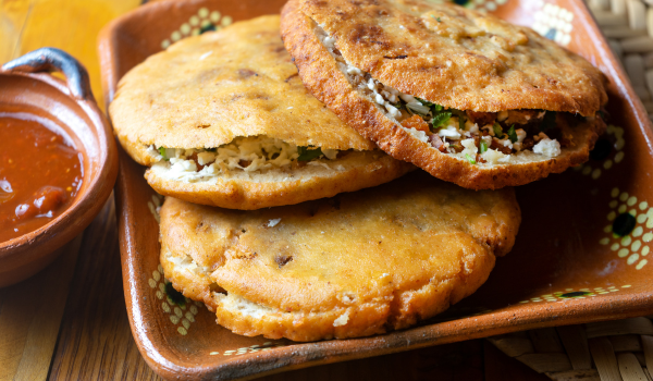 Golden-brown g with cheese, fresh herbs, and seasoned fillings, served in a traditional Mexican clay plate with a side of salsa. The rustic setup adds a touch of cultural authenticity