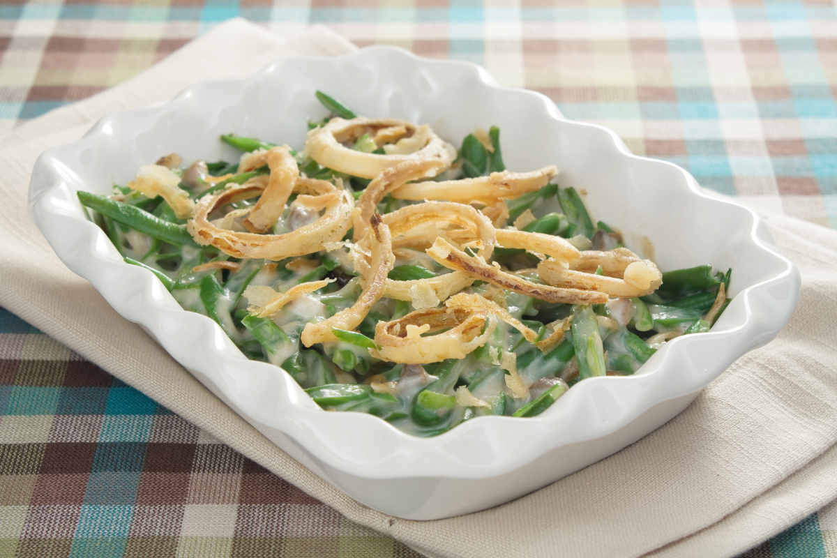 Close-up of a baked green bean casserole recipe in a white dish, topped with golden-brown fried onions on a plaid tablecloth.