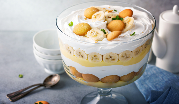 A stylish glass bowl filled with layers of vanilla pudding, fresh banana slices, vanilla wafers, and whipped cream, topped with cookies and fresh mint leaves—perfect for a classic and comforting dessert.