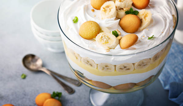 A close-up of homemade banana pudding in a glass bowl, showcasing visible layers of creamy pudding, banana slices, vanilla wafers, and a whipped cream topping garnished with cookies and fresh mint.