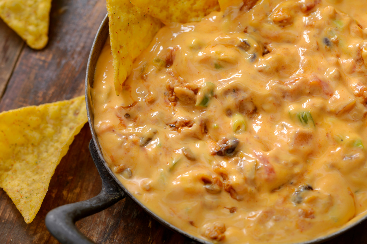 A close-up view of a thick, orange cheese dip filled with meat and chiles in a cast-iron skillet, surrounded by crunchy tortilla chips. rotel dip