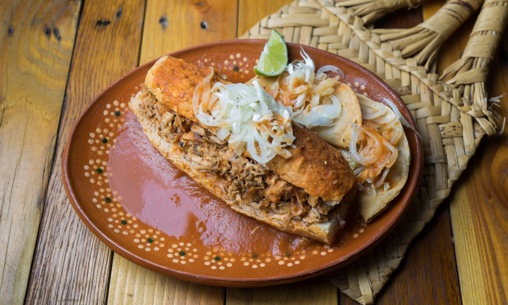 A delicious Mexican torta drenched in red sauce, topped with shredded cabbage, and served with crispy tacos on a colorful traditional Mexican plate. A perfect representation of authentic Mexican food.