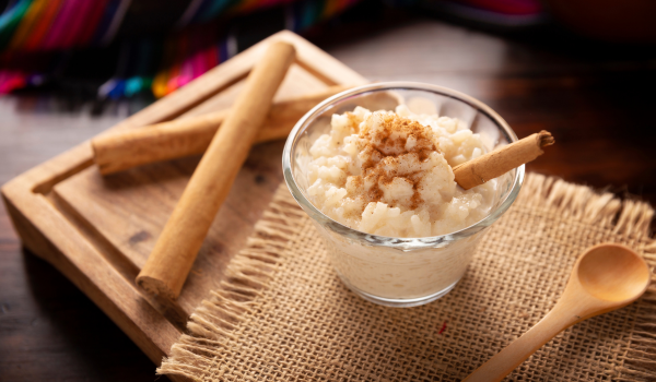 Creamy arroz con leche in a glass dish, topped with ground cinnamon and cinnamon sticks, accompanied by a wooden spoon on a jute base.