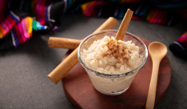 Traditional Mexican arroz con leche in a glass bowl, sprinkled with cinnamon, presented on a dark wooden board with cinnamon sticks in the background.