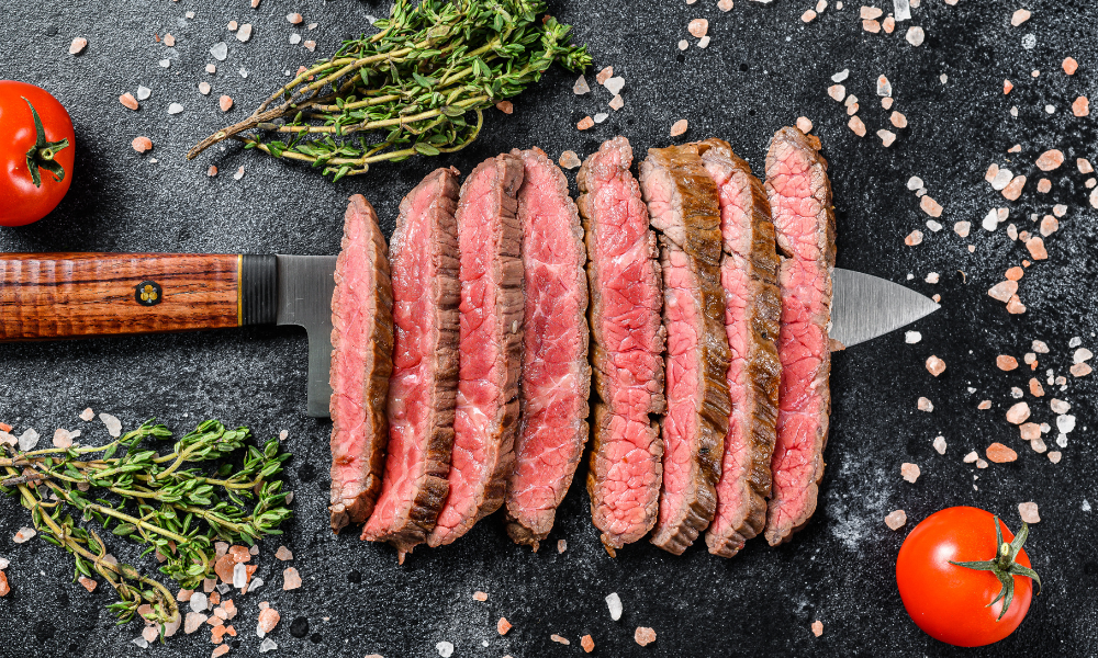 Authentic Carne Asada Recipe: the Best Carne Asada You’ll Ever Make! 12 Perfectly cooked carne asada slices arranged on a knife, surrounded by fresh herbs and tomatoes on a dark slate surface.