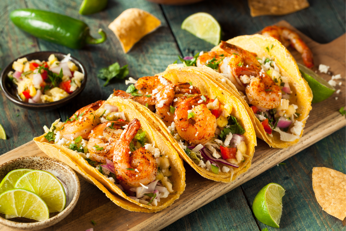 Golden tortillas filled with juicy shrimp, crisp lettuce, red onion, and a sprinkling of queso fresco. Lime wedges and fresh salsa complete the scene, highlighting bright flavors and a zesty kick.