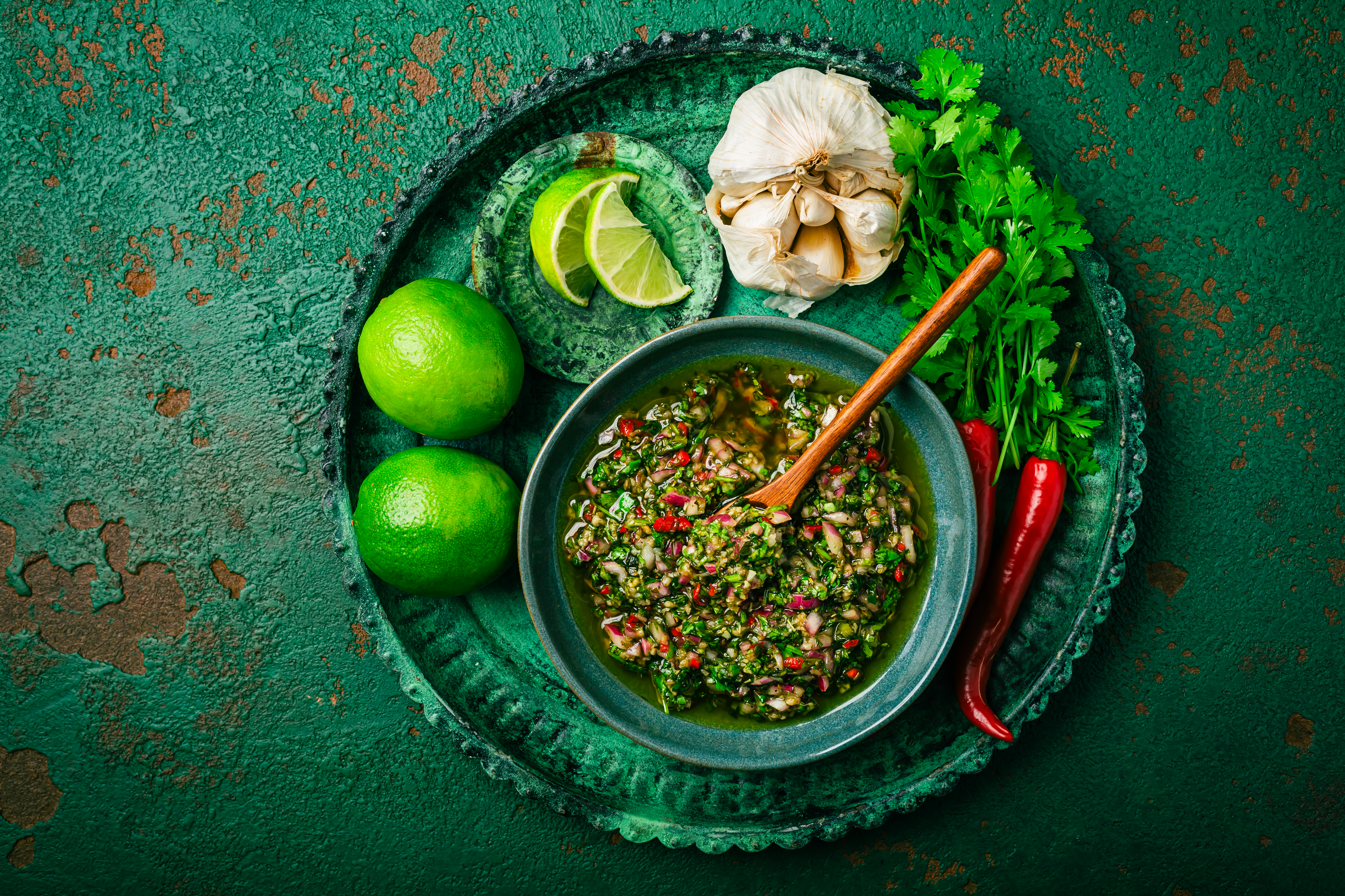 Homemade chimichurri sauce served in a bowl with fresh lime, parsley, garlic, and red chilies on a textured green surface