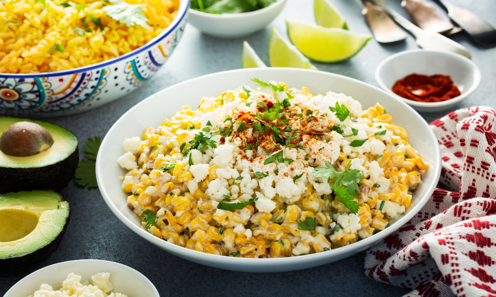 A vibrant plate of Mexican street corn salad (esquites) topped with cotija cheese, cilantro, and chili powder, surrounded by fresh avocado, lime wedges, and colorful garnishes