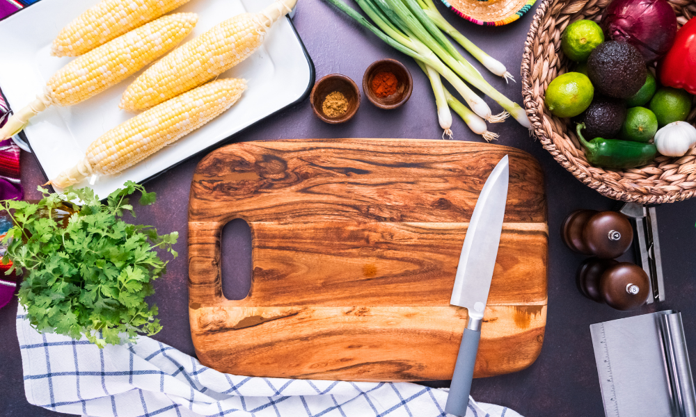 A beautifully arranged preparation area for Mexican street corn salad (esquites), featuring fresh corn, cilantro, green onions, spices, limes, avocados, and vibrant vegetables alongside a wooden cutting board and knife.