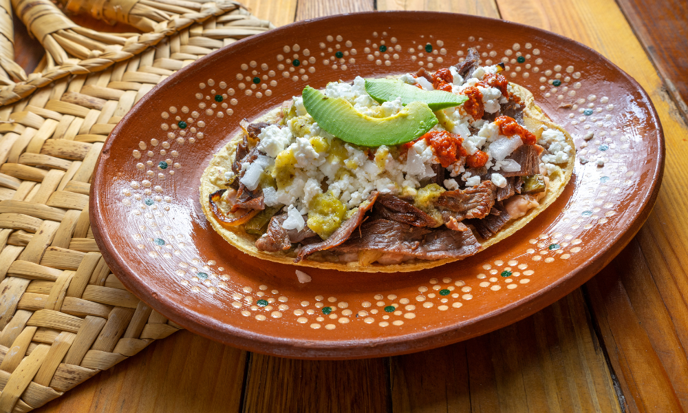 A classic Mexican huarache adorned with savory beef, creamy cheese, fresh avocado slices, and vibrant red and green salsas, served alongside small bowls of salsa for dipping.