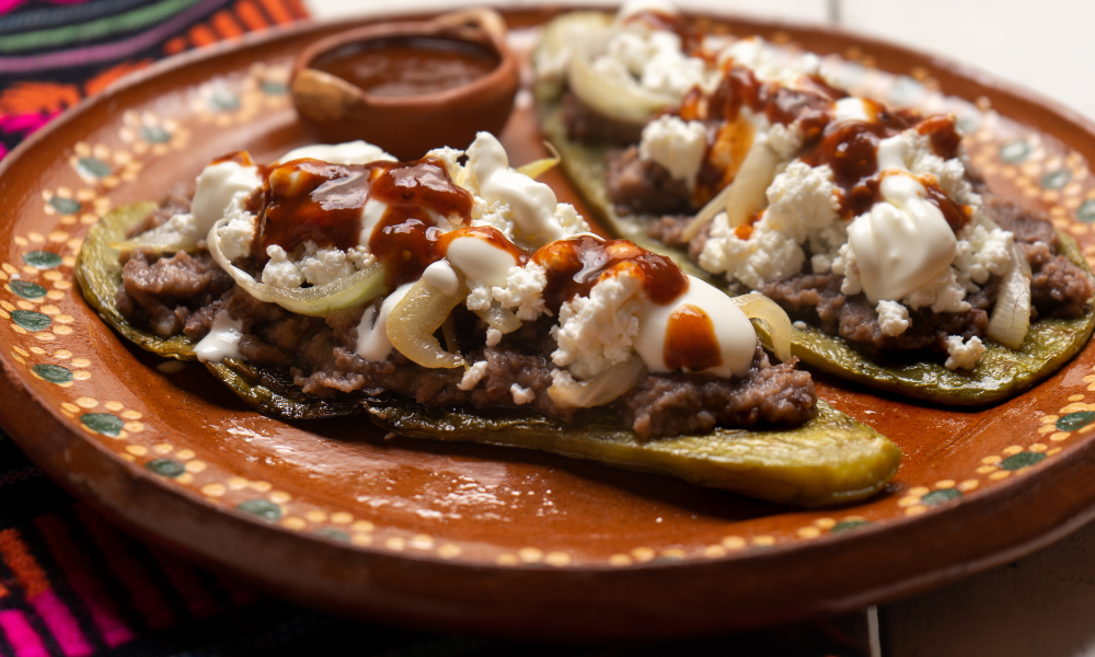 Nopal huaraches layered with refried beans, crumbled cheese, sour cream, caramelized onions, and a drizzle of spicy salsa, highlighting a vibrant and traditional Mexican dish