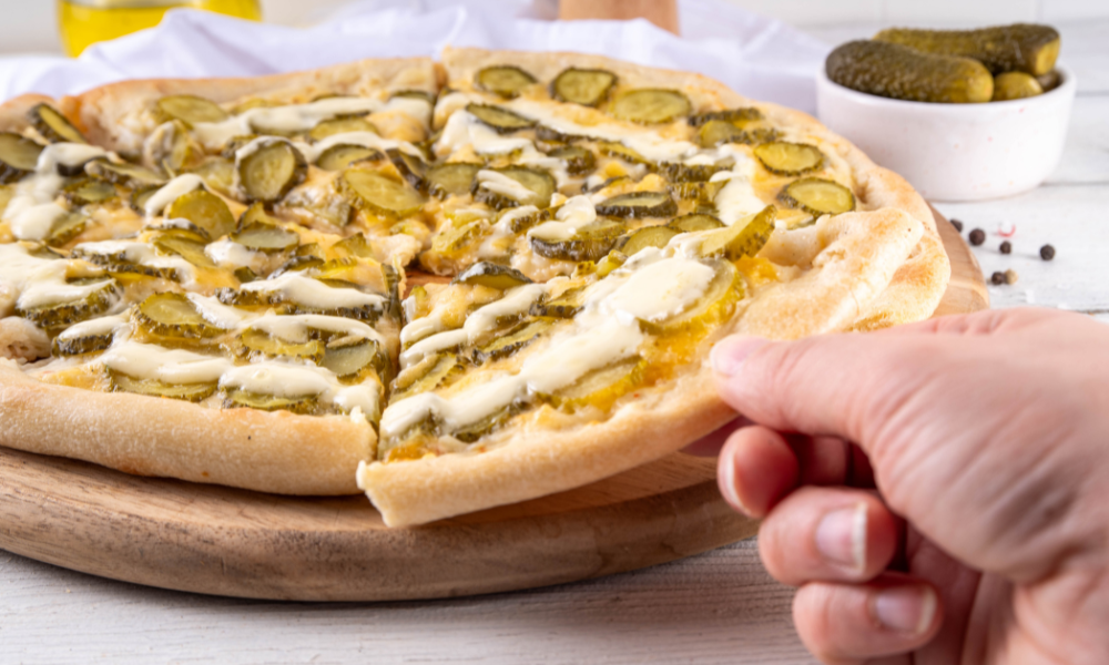 Pickle Pie Pizza Hand lifting a slice of pickle pizza with visible layers of pickles, cheese, and golden crust.