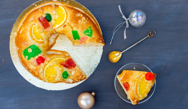 Close-up of a Three kins' cake with colorful candied fruits and almond slices on a golden base, with a slice served on a small plate beside festive ornaments.