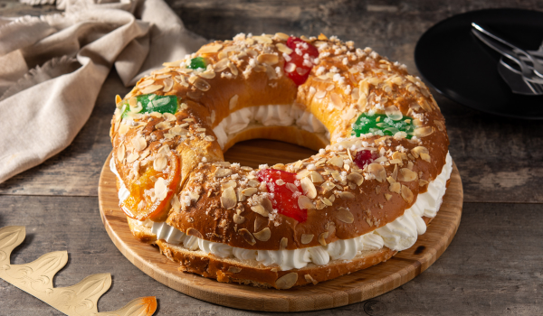 King cake filled with whipped cream, topped with candied fruits, almond slices, and powdered sugar on a wooden serving board.